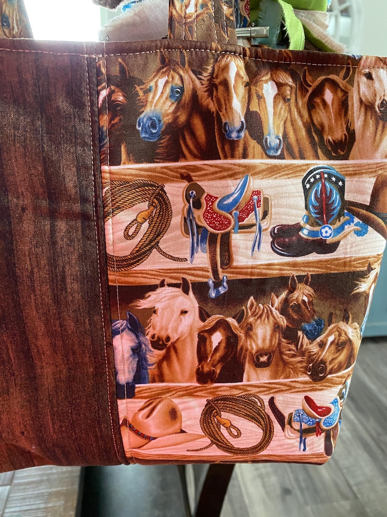 May include: A fabric tote bag featuring a repeating pattern of horses, western saddles, cowboy boots, lassos, and hats. The design includes a dark brown wood-grain textured side panel and a main section with horizontal rows of equestrian imagery.