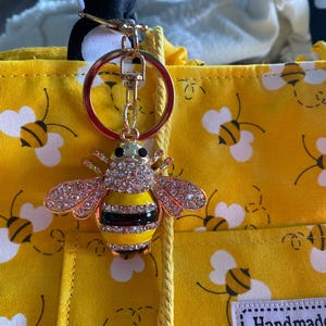 May include: A gold-toned metal bee keychain featuring clear rhinestones on the wings and thorax, with yellow and black enamel stripes on the abdomen. The keychain is attached to a yellow fabric bag patterned with small bees and white wings.