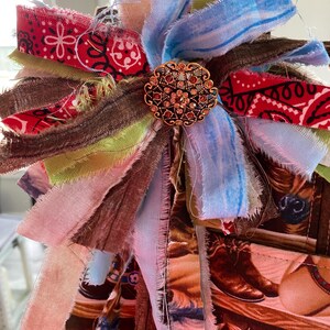 May include: A decorative bow made of layered, frayed fabric strips in red, blue, brown, and light green, featuring a central round gold-toned metal brooch with intricate filigree patterns and embedded small amber-colored rhinestones.