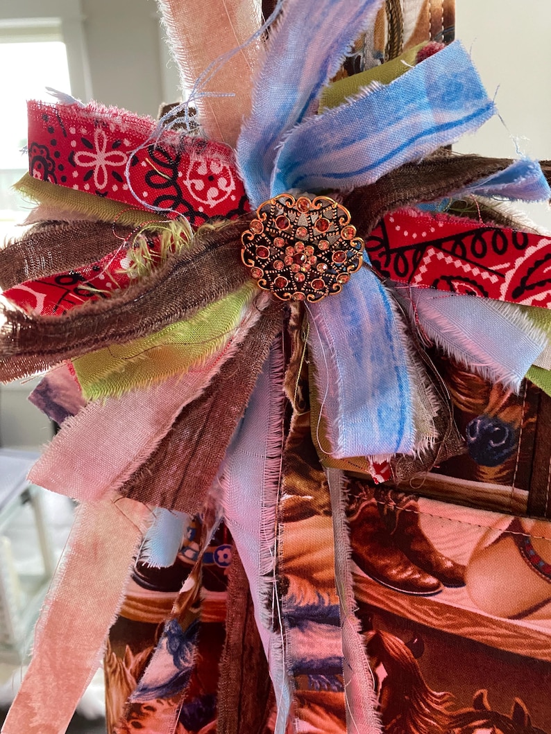 May include: A decorative bow made of layered, frayed fabric strips in red, blue, brown, and light green, featuring a central round gold-toned metal brooch with intricate filigree patterns and embedded small amber-colored rhinestones.