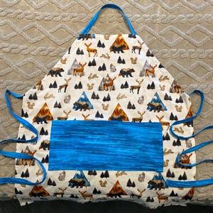 May include: A white apron with a blue pocket and ties. The apron is patterned with brown bears, deer, squirrels, mountains, and trees. The pocket has a blue, textured design. The apron has blue ties and a blue neck strap.