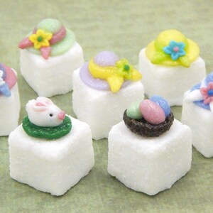 May include: Eight white sugar cubes decorated with Easter-themed fondant toppers. The toppers include a bunny rabbit, a nest with eggs, and various colored hats with flowers.