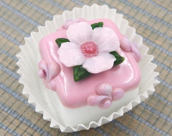 May include: A pink and white fondant cupcake with a delicate pink flower on top. The cupcake is in a white paper liner.