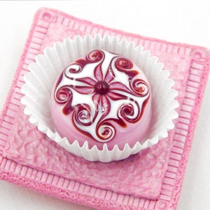 May include: A pink and white glass candy with a swirl design, sitting in a white paper cup on a pink background.