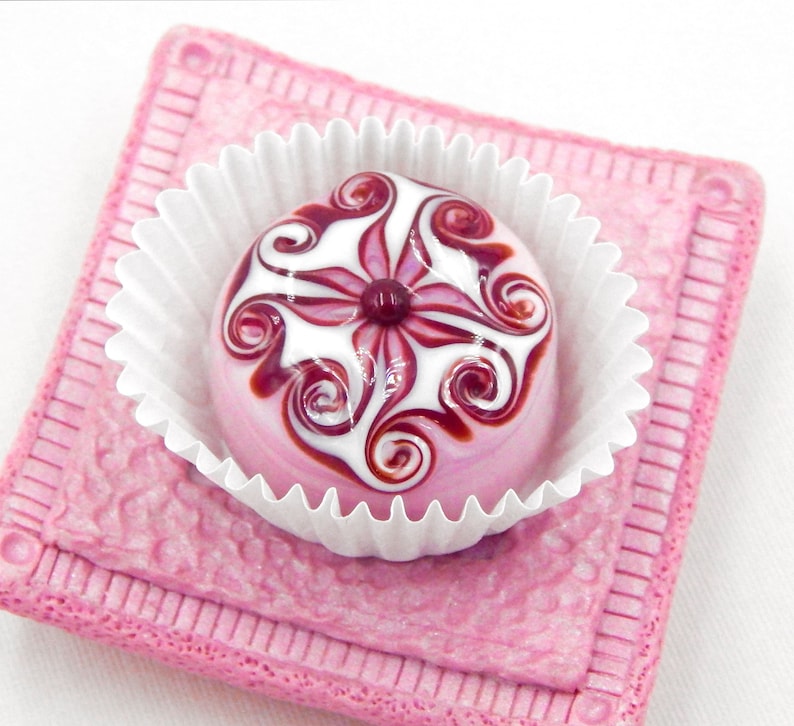 May include: A pink and white glass candy with a swirl design, sitting in a white paper cup on a pink background.