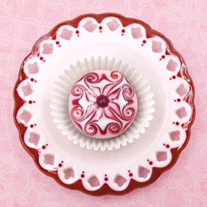 May include: A white and pink swirl design cupcake on a white and red plate with a scalloped edge.