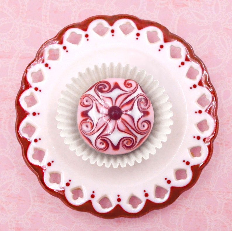 May include: A white and pink swirl design cupcake on a white and red plate with a scalloped edge.
