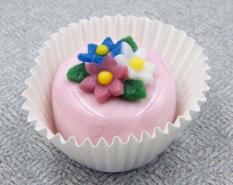 May include: A pink cupcake with white frosting and a decorative sugar flower arrangement. The flowers are blue, pink, white, and green.