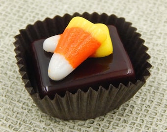 May include: A dark chocolate candy with a yellow, orange, and white candy corn on top. The candy is in a brown paper cupcake liner.