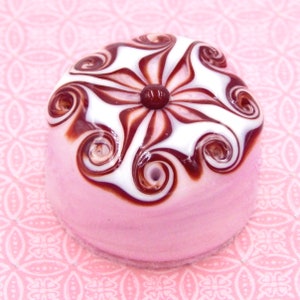 May include: A pink glass bead with a white and brown swirl design. The bead has a red center and is shaped like a dome.