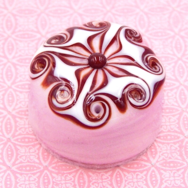 May include: A pink glass bead with a white and brown swirl design. The bead has a red center and is shaped like a dome.