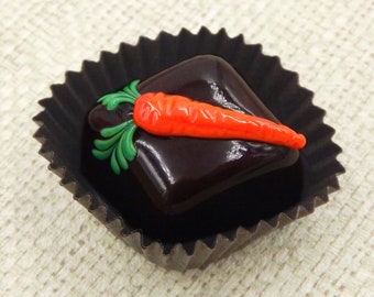 May include: A dark chocolate candy with a carrot-shaped decoration on top. The candy is in a black paper cup.