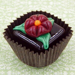 May include: A dark chocolate truffle topped with a red glass flower with a yellow center and green leaves, all in a brown paper cupcake liner.