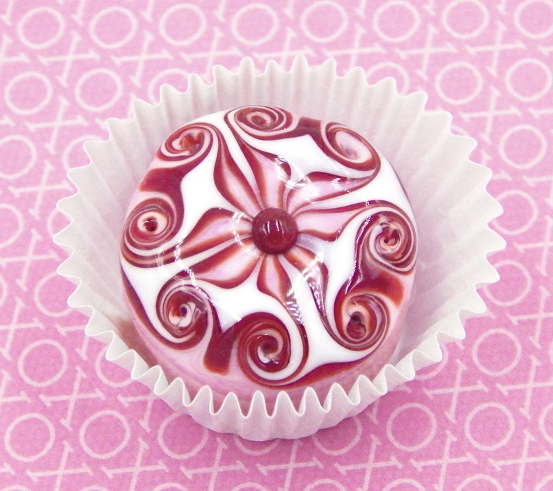 May include: A white and pink candy with a swirl design and a red center, sitting in a white paper cupcake liner.