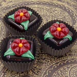 May include: Three chocolate truffles with red flower decorations in black paper cups on a gold patterned surface.