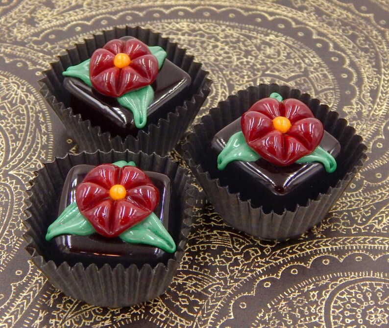 May include: Three chocolate truffles with red flower decorations in black paper cups on a gold patterned surface.