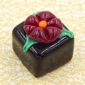 May include: A dark brown glass cube with a red glass flower on top. The flower has a green stem and leaves and an orange center.