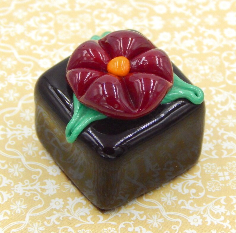 May include: A dark brown glass cube with a red glass flower on top. The flower has a green stem and leaves and an orange center.