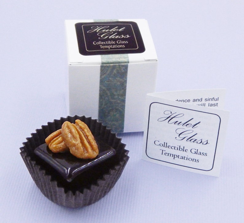 Puede incluir: Una peque&ntilde;a taza de papel marr&oacute;n oscuro llena de un cuadrado de chocolate cubierto con dos mitades de nuez pecana. La taza est&aacute; sobre una superficie blanca. El texto "Hulet Glass Collectible Glass Temptations" est&aacute; impreso en una etiqueta blanca con borde negro.