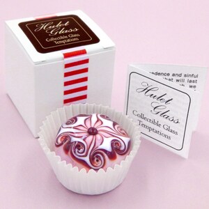 May include: A small, white box with a red and white striped ribbon. The box is labeled "Hulet Glass Collectible Glass Temptations". A small, pink and white glass paperweight with a swirl design is sitting in a white paper cup. The paperweight is also labeled "Hulet Glass Collectible Glass Temptations".