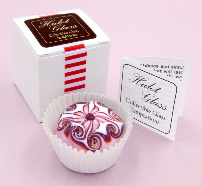 May include: A small, white box with a red and white striped ribbon. The box is labeled "Hulet Glass Collectible Glass Temptations". A small, pink and white glass paperweight with a swirl design is sitting in a white paper cup. The paperweight is also labeled "Hulet Glass Collectible Glass Temptations".