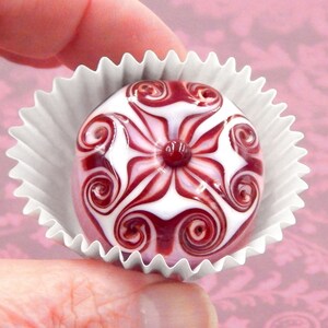 May include: A white and burgundy glass bead with a swirling floral design. The bead is in a white paper cupcake liner.