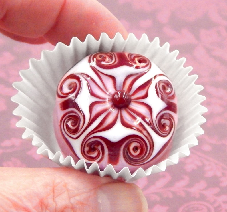 May include: A white and burgundy glass bead with a swirling floral design. The bead is in a white paper cupcake liner.