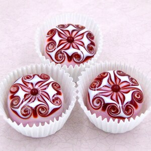 May include: Three pink glass ornaments with white and red swirling designs. Each ornament is in a white paper cup.