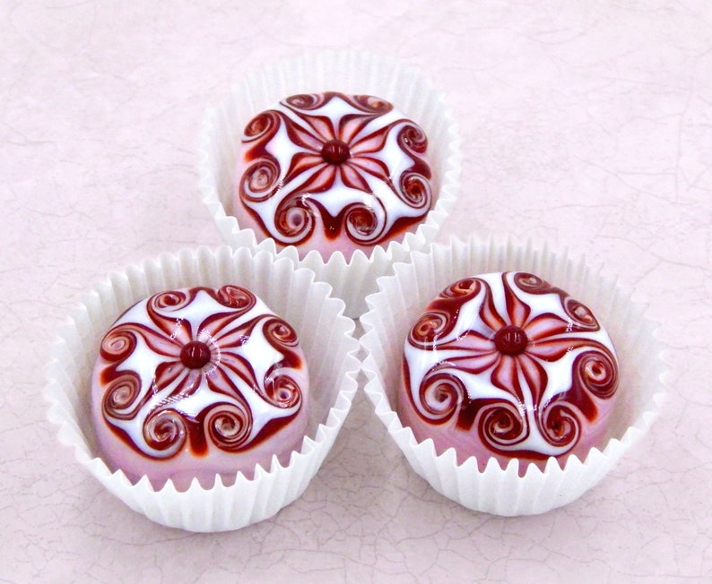 May include: Three pink glass ornaments with white and red swirling designs. Each ornament is in a white paper cup.
