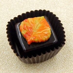 May include: A dark chocolate truffle with a decorative orange and yellow leaf design on top. The truffle is in a black paper cup.