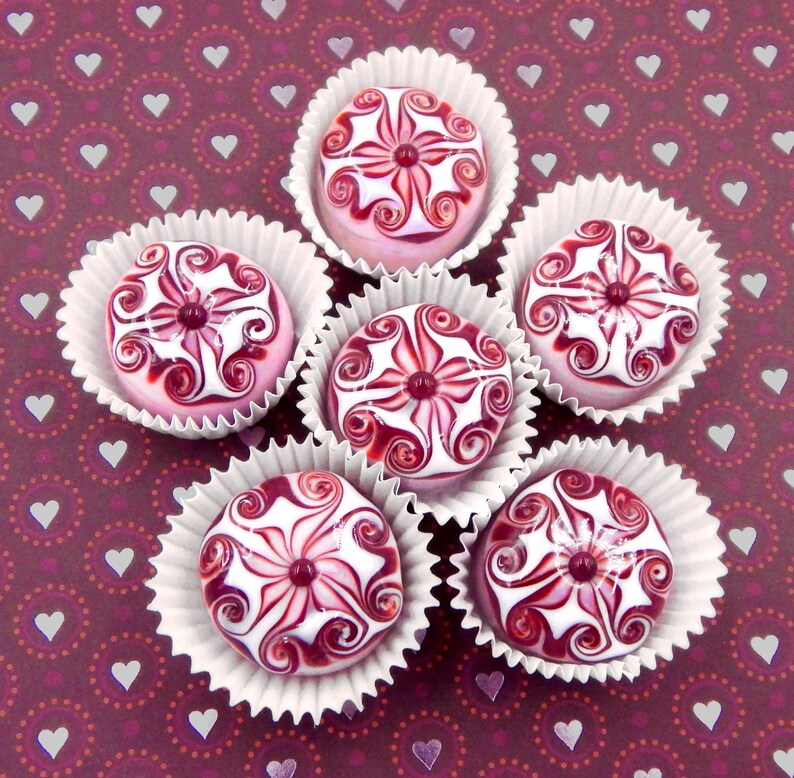May include: Six white cupcake liners with pink and white swirl designs. Each cupcake liner contains a pink and white swirl candy. The candies are arranged in a circle on a burgundy background with white hearts.