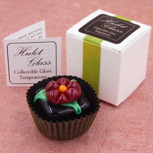 May include: A small glass flower in a dark brown chocolate-like cup. The flower is red with a yellow center and green leaves. The cup is sitting on a pink surface. The text on the box reads "Hulet Glass Collectible Glass Temptations".