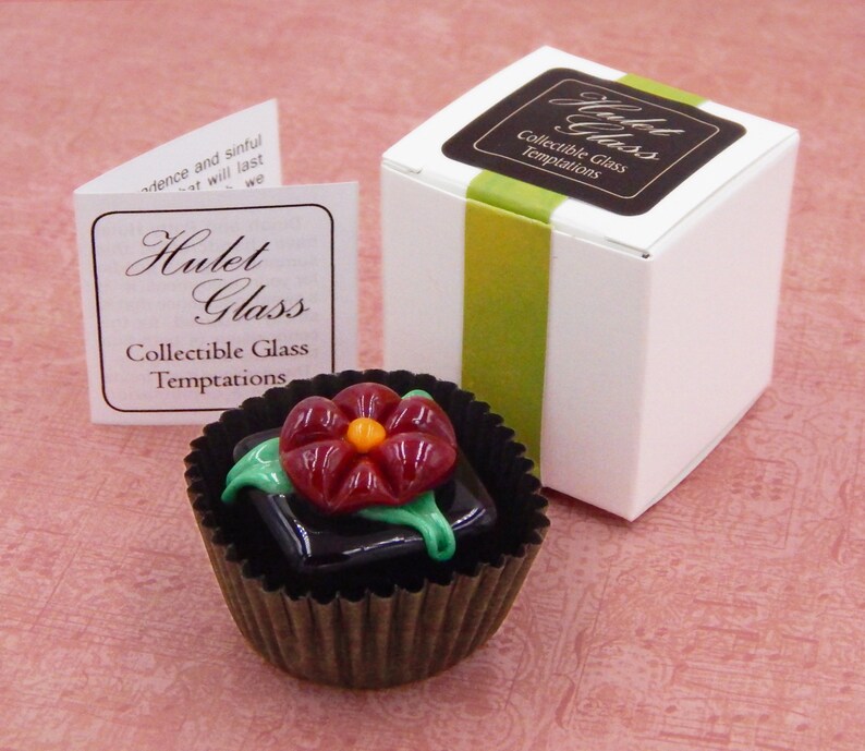 May include: A small glass flower in a dark brown chocolate-like cup. The flower is red with a yellow center and green leaves. The cup is sitting on a pink surface. The text on the box reads "Hulet Glass Collectible Glass Temptations".