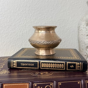 May include: A small, antique brass vase with a decorative, textured band around the middle. The vase sits atop a vintage book with the title "Tennyson" in gold lettering. The book is on top of another book with gold accents.