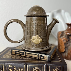May include: A brown ceramic teapot with a rounded lid and a circular handle. The teapot has a decorative design on the front and sits atop two antique books. A small, brown vase is visible in the background.