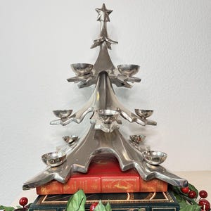 May include: A silver-colored, tree-shaped candle holder with multiple small bowl-shaped candle cups. The candle holder sits atop two antique books with red and green covers. A garland of red berries and green leaves decorates the base.