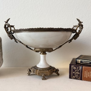 May include: An antique white and gold decorative bowl with ornate handles and a pedestal base. The bowl has a white interior and a detailed rim. The base is a detailed gold color. The bowl is a decorative item.