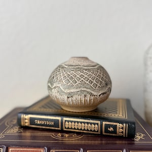 Może przedstawiać: Mały, okrągły, beżowo-szary wazon ceramiczny z teksturowanym, żłobionym wzorem kratki i falistą linią poziomą. Wazon stoi na czarnej książce z napisem Tennyson, która spoczywa na większej, ciemnobordowej książce ze złotymi literami na okładce.