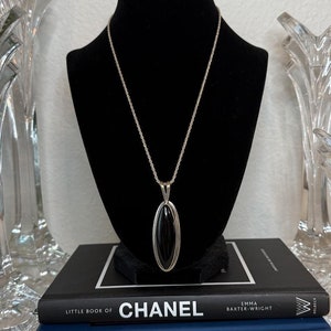 May include: A silver chain necklace with a black oval pendant. The necklace is displayed on a black mannequin bust. The bust is sitting on top of two books, one with the title "Little Book of Chanel" and the other with the title "Little Book of Alexander McQueen".