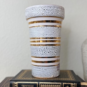 May include: A white and gold ceramic vase with a textured surface and horizontal gold stripes. The vase is cylindrical and sits atop a vintage book with the title "Tennyson" in gold lettering.