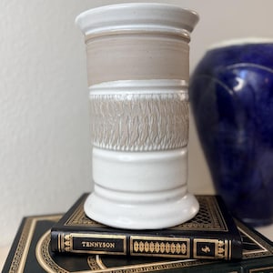 Può includere: Un vaso in ceramica fatto a mano con una fascia strutturata marrone chiaro e dettagli bianchi. Il vaso è posto su una pila di libri, uno dei quali intitolato "TENNYSON". Un vaso blu è sullo sfondo.