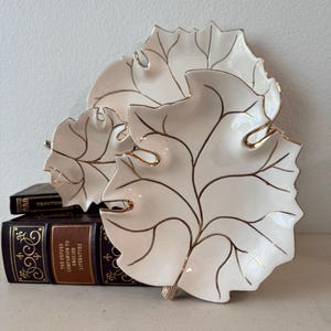 May include: Three cream-colored ceramic dishes shaped like leaves, each featuring delicate gold-painted veins and edges. The dishes are arranged in a tiered stack, with the largest at the back and two smaller ones in front, resting against a book.