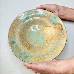 May include: A round ceramic bowl featuring a crystalline glaze with a blend of light blue, turquoise, and warm tan tones. The surface has a glossy finish with organic, mottled patterns that transition between the blue and earthy brown colors.