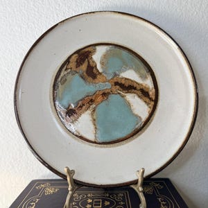 Könnte beinhalten: Ein Keramikteller mit cremefarbenem Rand und einem zentralen Design mit abstrakten blauen, braunen und weißen Formen. Der Teller hat einen dunkelbraunen Rand und wird auf einem dekorativen Metallständer präsentiert. Es ist ein dekorativer Artikel.