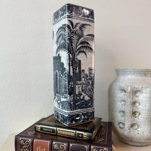 May include: A tall, square ceramic vase featuring a black and white toile pattern with palm trees and architectural structures. The surface has a visible crackle glaze finish. It is positioned atop two stacked hardcover books.