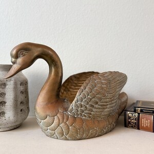 May include: A bronze-colored swan figurine with detailed wing and feather patterns. The swan has a curved neck and is positioned on a white surface. A textured vase and a stack of books are also present.
