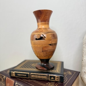 May include: A decorative vase constructed from various small, rectangular pieces of light and dark brown wood. The vase features a flared rim, a rounded body with a visible natural knot hole, and a dark brown circular base.