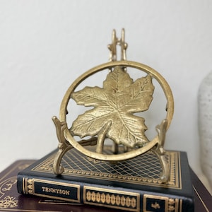 May include: A brass-colored book stand featuring a maple leaf design within a circular frame. The stand is positioned on top of a vintage book with the title "Tennyson" in gold lettering. The book has a black cover with gold accents.