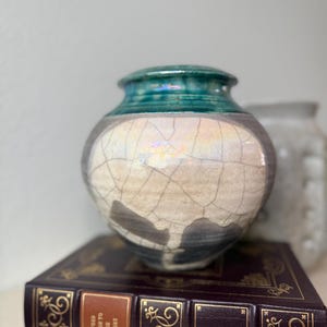 Pode incluir: Um vaso de cerâmica com uma borda verde-azulada brilhante e um corpo esbranquiçado perolado e rachado com detalhes em cinza escuro na base. O vaso repousa sobre um livro de capa de couro marrom escuro intitulado The Oxford Companion to English Literature.