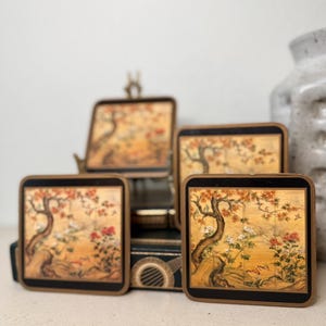 May include: Set of four square coasters with a vintage Asian landscape design. Each coaster features a detailed illustration of a tree, flowers, and birds in shades of orange, red, and gold, framed with a black border.
